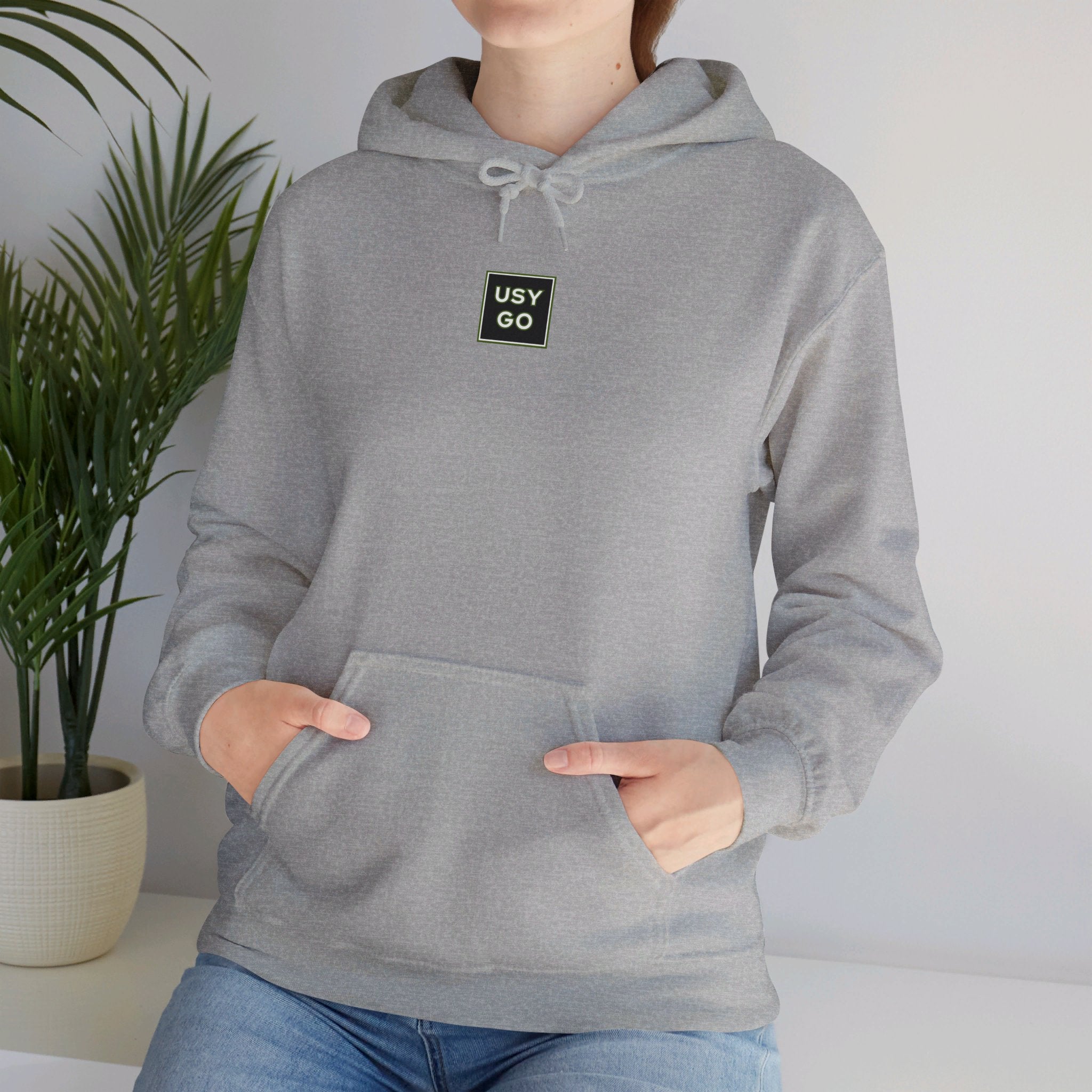 Person wearing a Heavy Blend Hoodie Gildan 18500 Unisex Jersey with USYGO logo on the front. Made from 50% Cotton 50% Polyester in Sport Grey. Pouch pocket and the tear-away label make for a highly comfortable, scratch-free wearing.