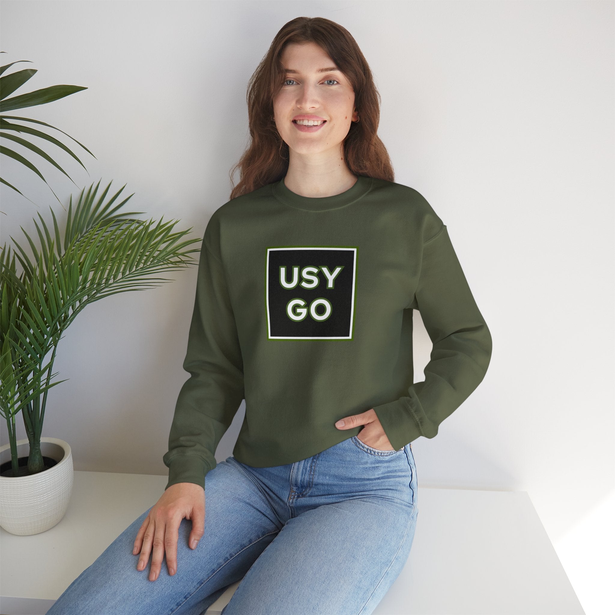 Person wearing a Heavy Blend Crewneck Sweatshirt Gildan 18000 Unisex with USYGO logo on the front. Made from 50% Cotton 50% Polyester in Military Green.