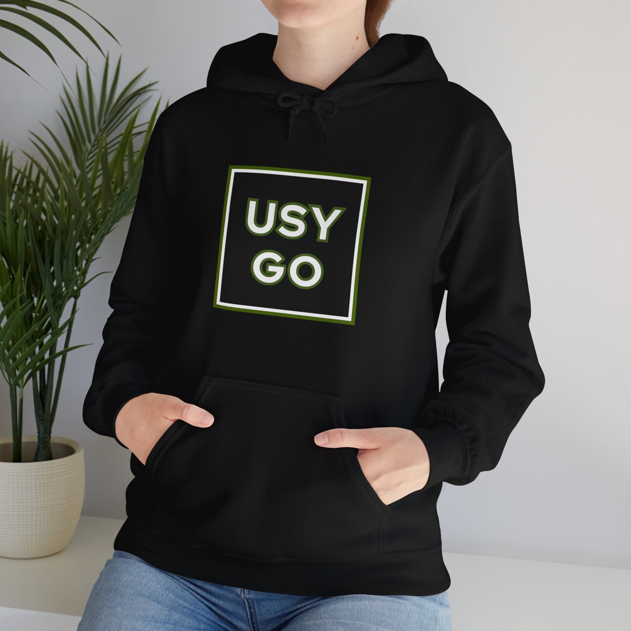 Person wearing a Heavy Blend Hoodie Gildan 18500 Unisex Jersey with big USYGO logo on the front. Made from 50% Cotton 50% Polyester in Black. Pouch pocket and the tear-away label make for a highly comfortable, scratch-free wearing.