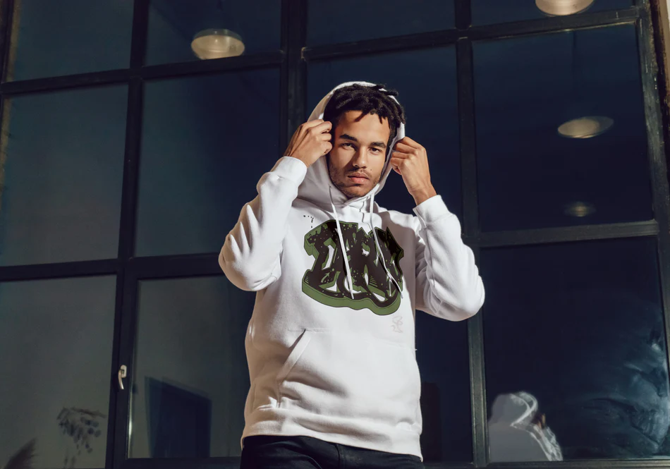 Young man adjusting the hood of his white streetwear hoodie featuring a bold graffiti-style graphic, standing indoors in front of large industrial windows.
